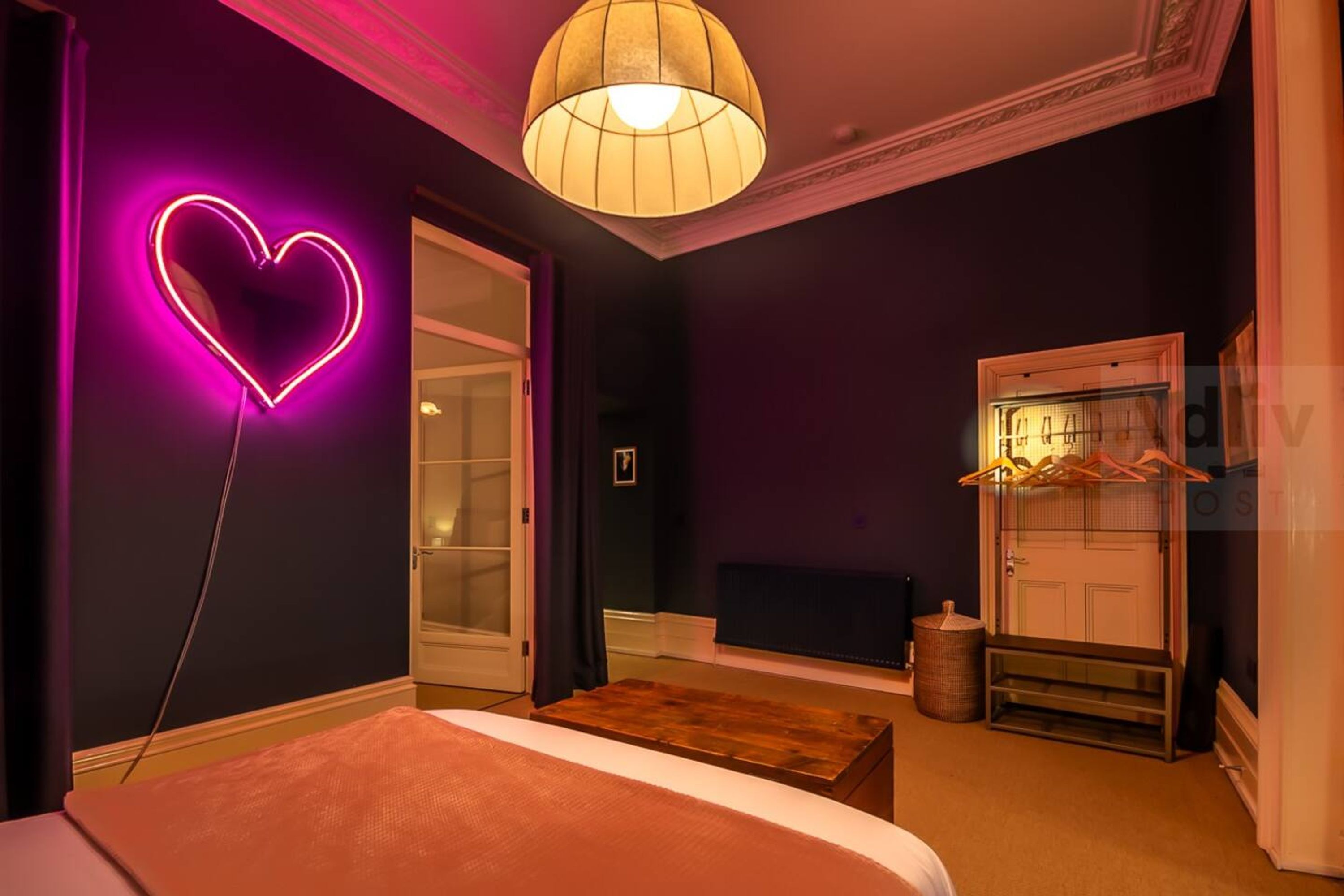 A room with a bed, a wooden bench, a neon heart sign, a hanging light fixture, and a door with a coat rack.