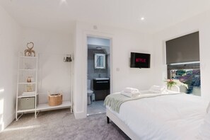 A modern hotel room with a neatly made bed, a TV mounted on the wall, a bookshelf, and a view of the outside through a window.