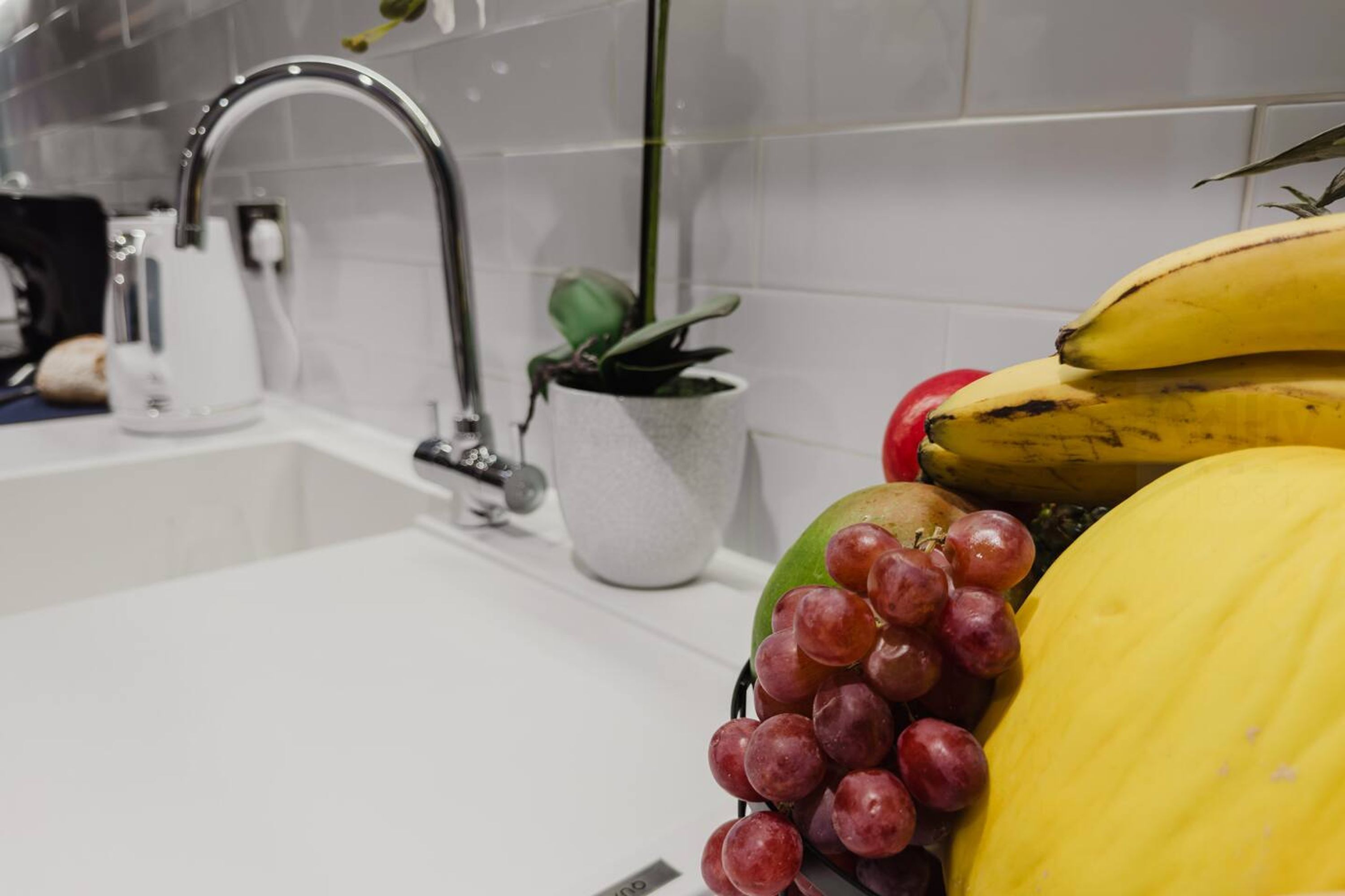 En vask med hane, en potteplante og en skål med bananer, druer og en melon.