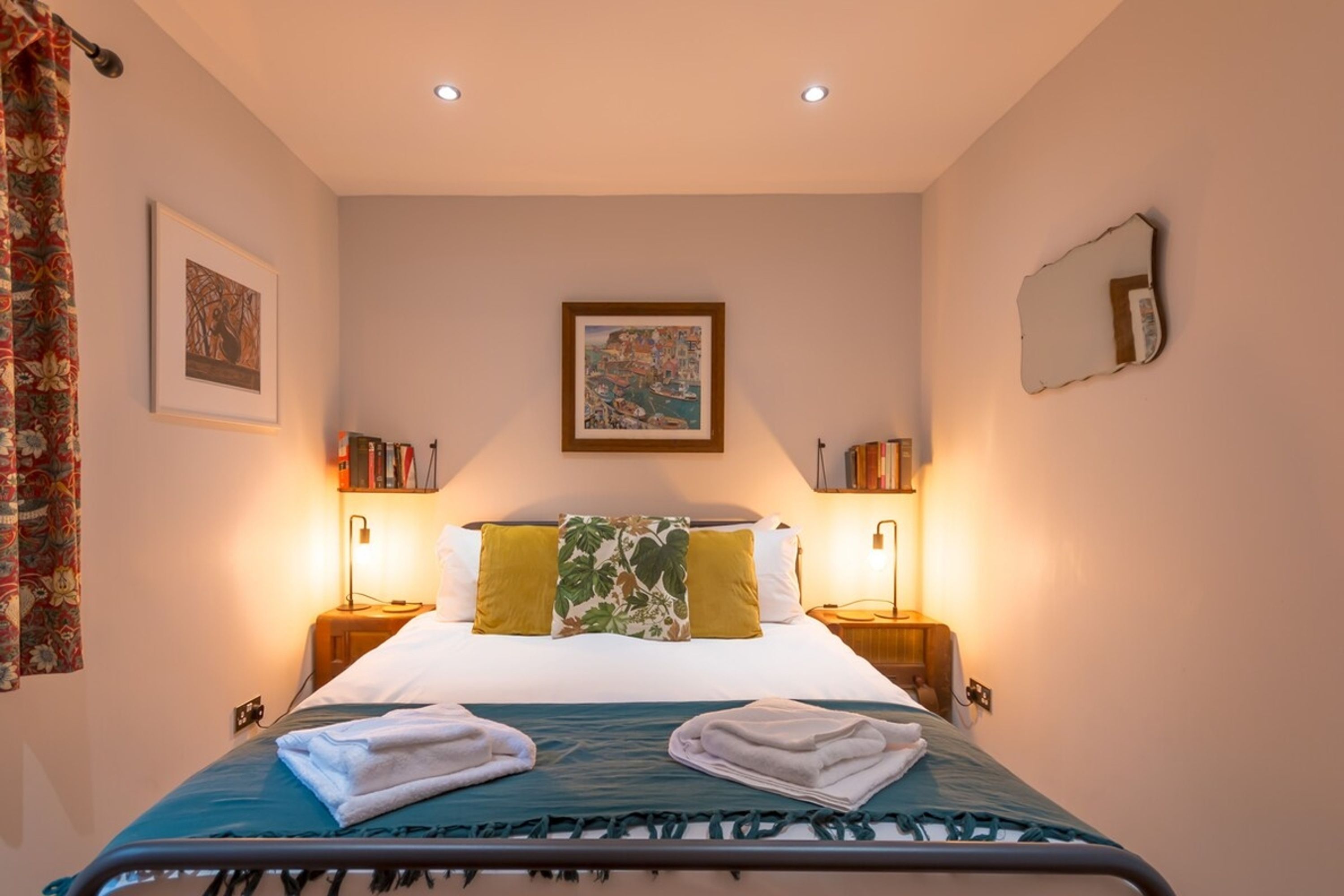 A neatly made bed with a green and white bedspread, two pillows, and folded towels on a headboard. There are two bedside tables with lamps and books.