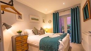 Ein ordentlich eingerichtetes Schlafzimmer mit Bett, Nachttischlampe, Kommode und Blick ins Freie durch eine Glastür.