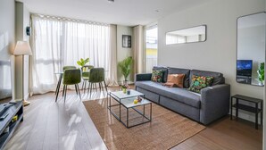 A modern living room with a grey sofa, a glass coffee table, and a dining area with green chairs. - Central London Stay in Vauxhall With Balcony (London)