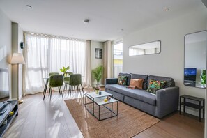 A modern living room with a grey sofa, a glass coffee table, and a dining area with green chairs. - Central London Stay in Vauxhall With Balcony (London)