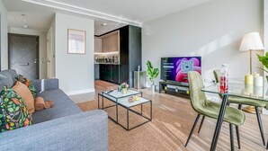 Un soggiorno moderno con un divano grigio, un tavolino da caffè in vetro e un'area pranzo con un tavolo in vetro e sedie verdi. Una TV è montata a parete, e sullo sfondo si vede una cucina.