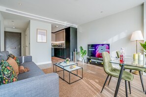 A modern living room with a grey sofa, a glass coffee table, and a dining area with a glass table and green chairs. A TV is mounted on the wall, and a kitchen is visible in the background. - Central London Stay in Vauxhall With Balcony (London)