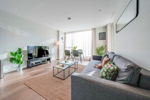 A modern living room with a grey sofa, a flat-screen TV, a coffee table, and a dining area with chairs and a table. - Central London Stay in Vauxhall With Balcony (London)