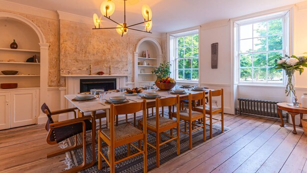 A dining room with a wooden table set for six, surrounded by chairs. A chandelier hangs from the ceiling, and there is a fireplace with a mantel. Shelves line the walls, and a window provides a view of greenery outside.