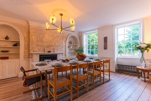 A dining room with a wooden table set for six, surrounded by chairs. A chandelier hangs from the ceiling, and there is a fireplace with a mantel. Shelves line the walls, and a window provides a view of greenery outside.