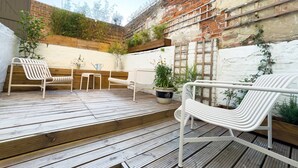 A wooden deck with white metal chairs and a small table, surrounded by brick walls and greenery.