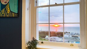 Een kamer met een groot raam met uitzicht op een zonsondergang, een potplant en een abstract kunstwerk aan de muur.