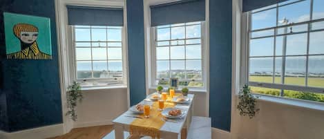 A dining area with a table set for breakfast, large windows with ocean views, and a painting on the wall.