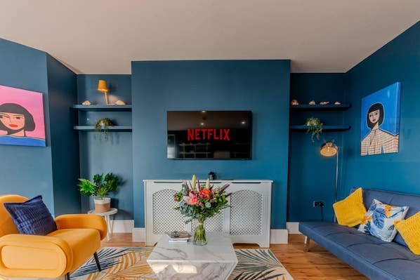 A modern living room with a blue accent wall, a Netflix sign, a sofa, a coffee table, and a TV. - Stunning Sea View Georgian Retreat by Adliv (Margate)