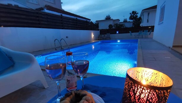 A poolside setting with a table, two glasses of water, a bottle, a plate with a food item, and a lit decorative lamp. - Sobella Villa - 3 Bedroom Villa With 10 x 4 Pool (Paralimni)