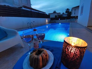 Une scène au bord de la piscine avec une table, deux verres d’eau, une bouteille, une assiette contenant un plat et une lampe décorative allumée.
