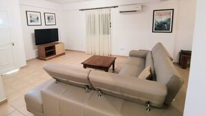 A living room with a sofa, a coffee table, a TV, and an air conditioning unit. - Sobella Villa - 3 Bedroom Villa With 10 x 4 Pool (Paralimni)