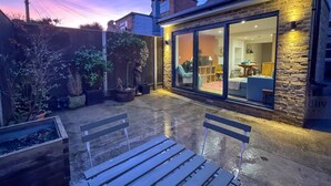 A patio with two wooden benches, a stone wall, and a glass door leading to a room with a sofa and a table. - Modern Margate Maisonette Close to Beach by Adliv (Margate)