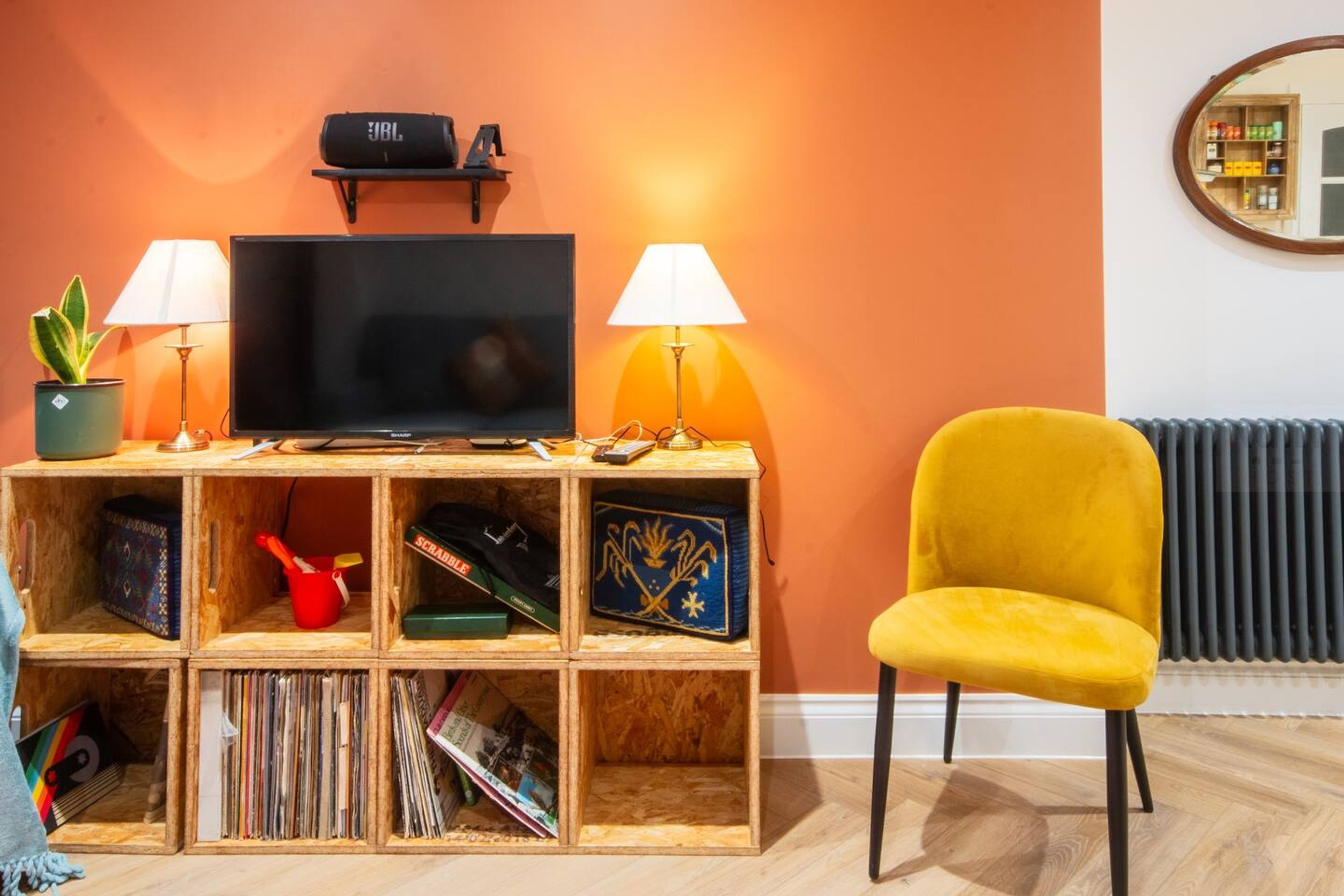A living room with a TV, two lamps, a yellow chair, a wooden shelf with items, and a radiator.