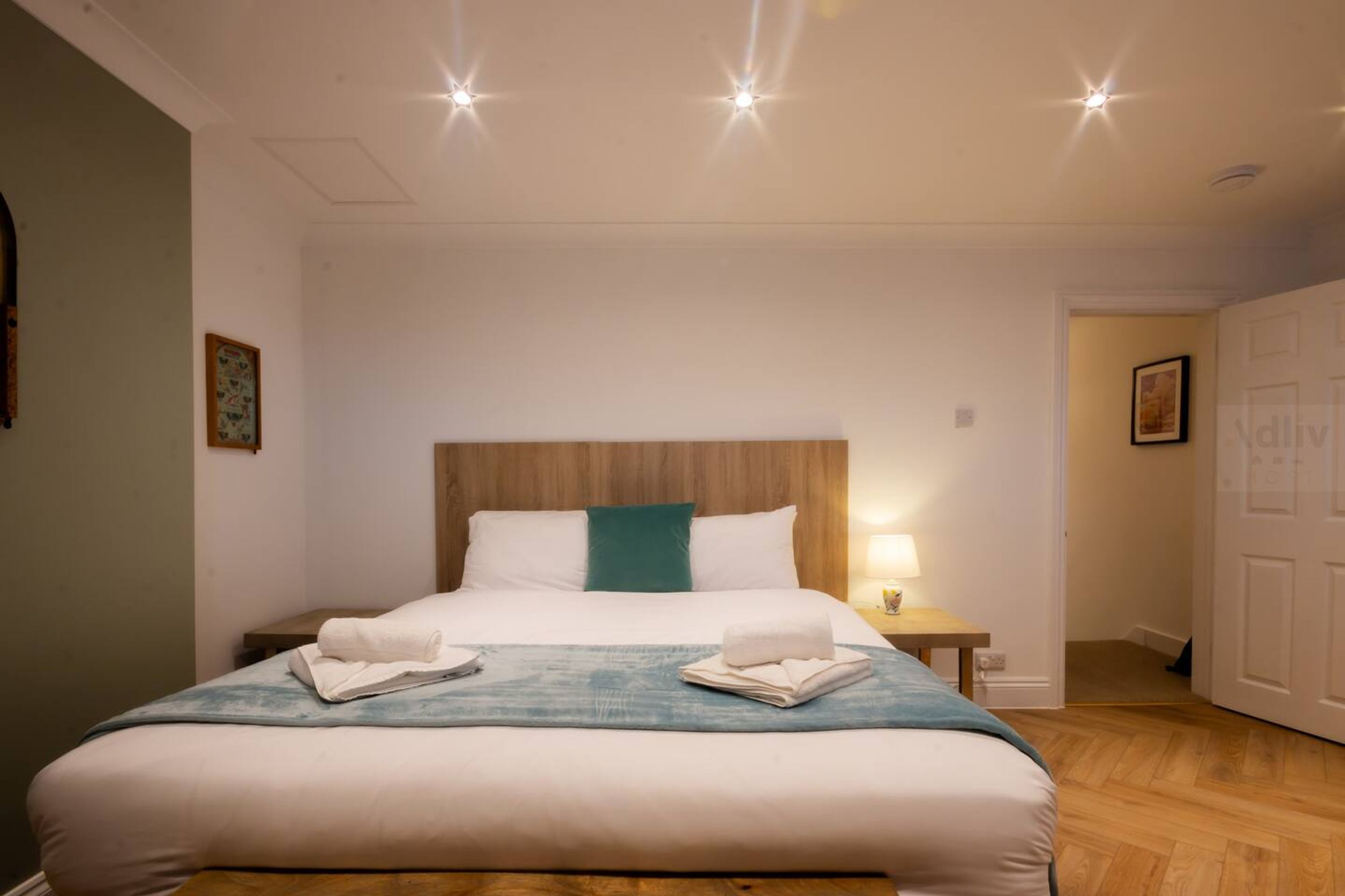 Una cama bien hecha con sábanas blancas y una manta color turquesa, un cabecero de madera y una mesita de noche con una lámpara.