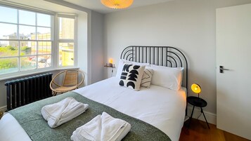Een net opgemaakt bed met witte lakens en een groene deken, een rieten stoel, een nachtkastje met een lamp, en een raam met uitzicht op gebouwen en groen.
