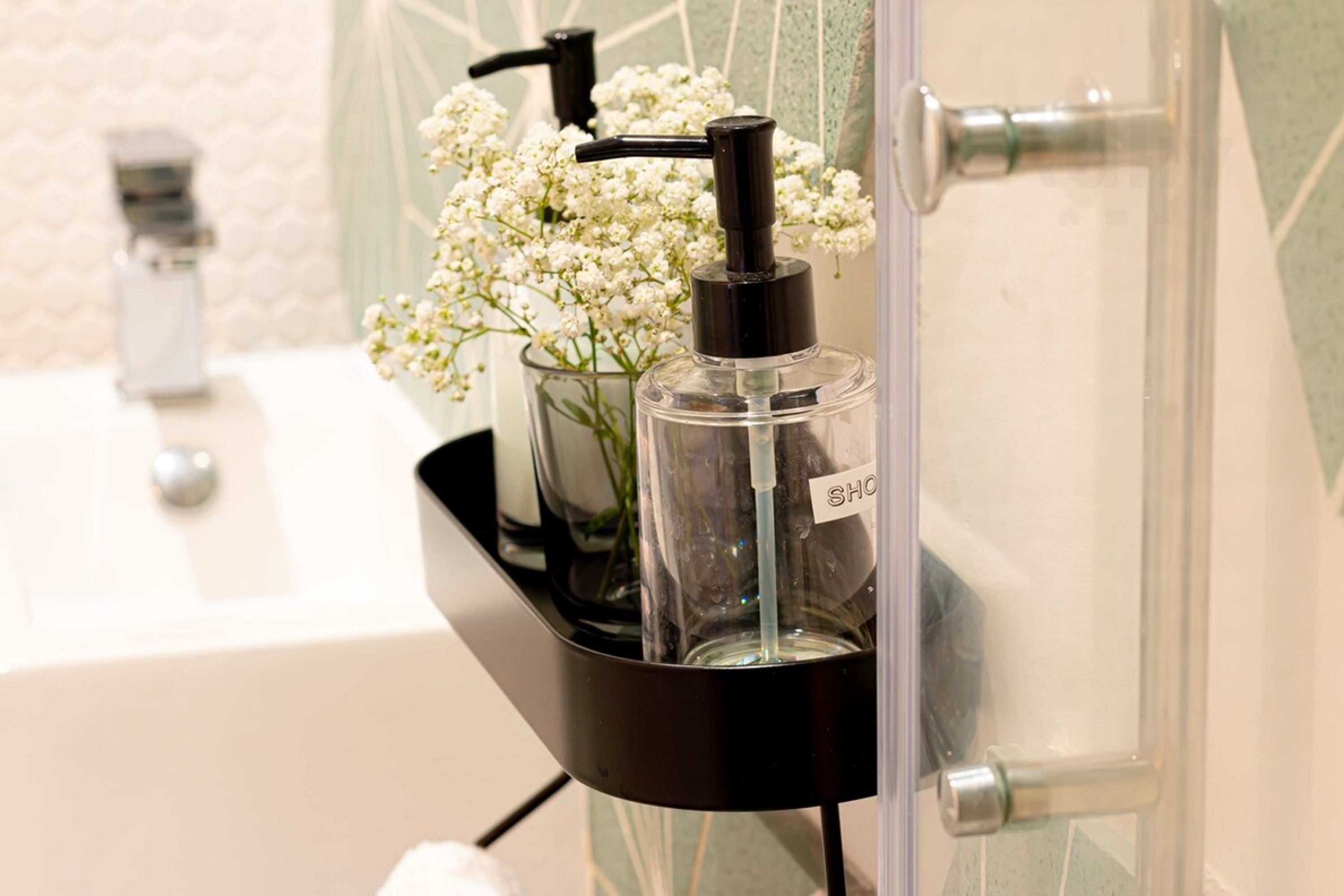 A bathroom with a black soap dispenser on a shelf, a clear glass container with flowers, and a white sink.