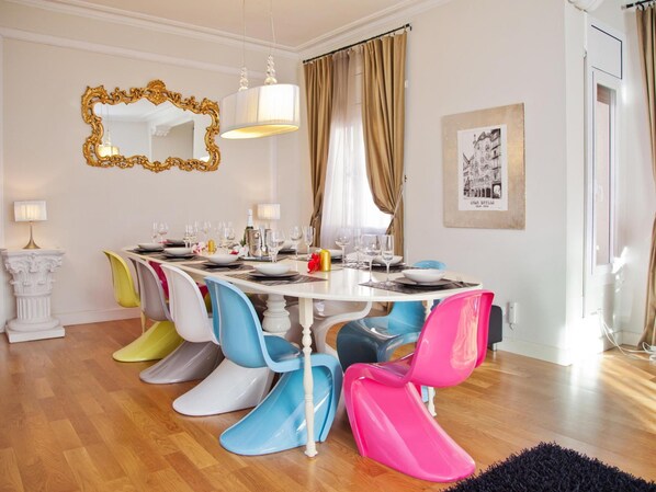 A dining area with a white table set for six, colorful chairs, a gold-framed mirror, and a framed picture on the wall.