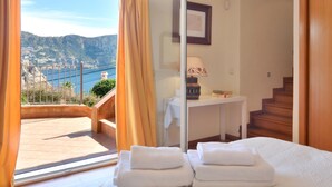 A hotel room with a large window, a bed with neatly folded white towels, a bedside table with a lamp, and a view of a coastal landscape. - Villa With Incredible sea Views and Pool Sleeps 7 (Andraitx)