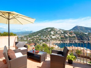 Une terrasse meublée de mobilier en osier, une table avec une bouteille et des fruits, et une vue sur une ville côtière.