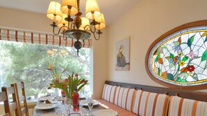 A dining area with a wooden table set for four, a bench with striped cushions, a chandelier, a stained glass window, and a painting on the wall. - Villa With Incredible sea Views and Pool Sleeps 7 (Andraitx)