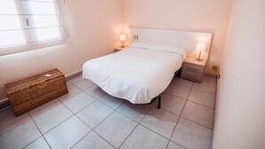 Een keurig opgemaakt slaapkamer met een bed, twee nachtkastjes met lampen, een klein rieten mandje en een raam met dunne gordijnen.