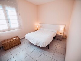 A neatly arranged bedroom with a bed, two bedside tables with lamps, a small wicker basket, and a window with sheer curtains.
