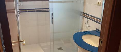Un baño con cabina de ducha de vidrio, un lavabo azul y un taburete de madera.