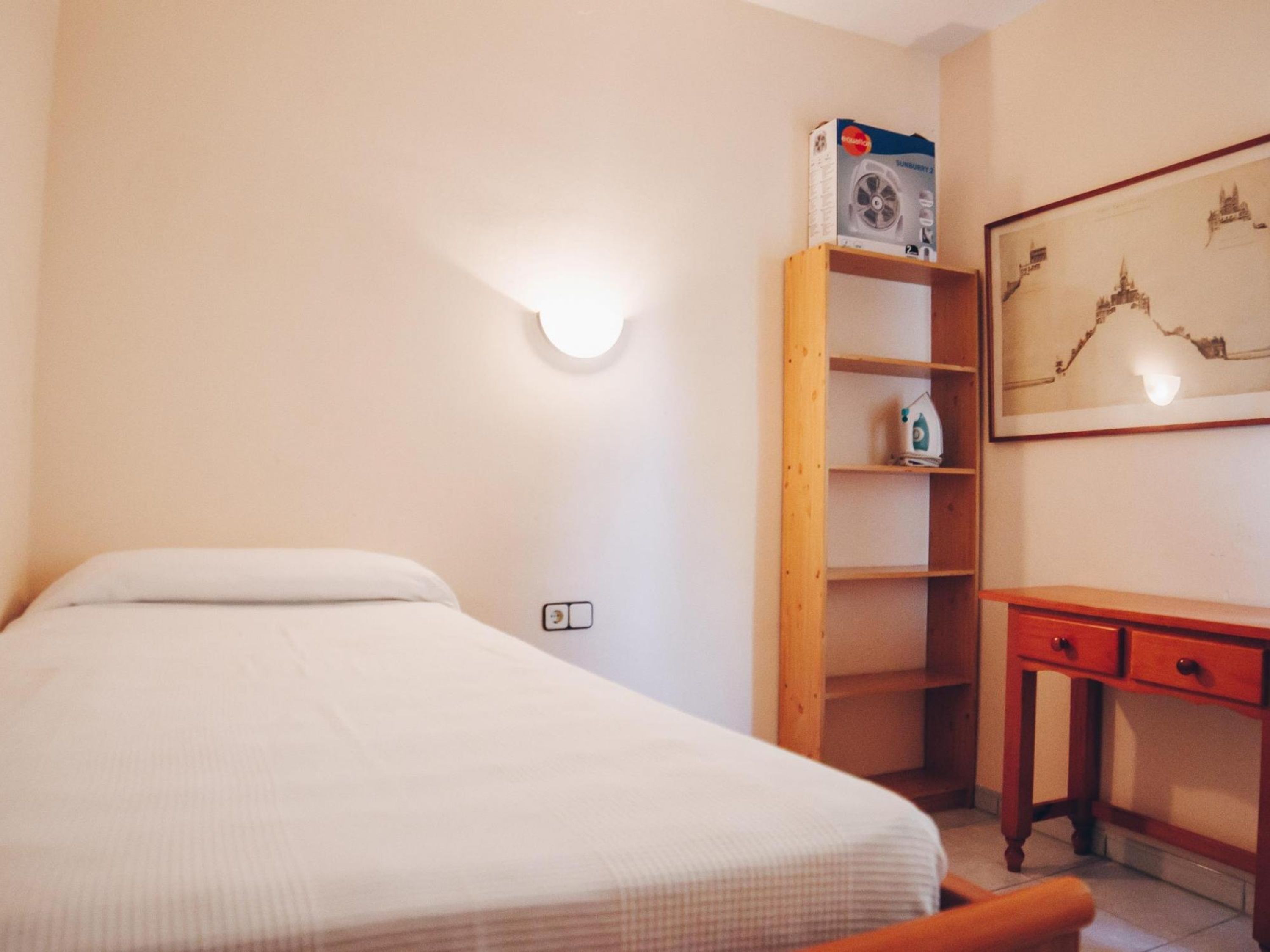 Une chambre à coucher avec un lit, une étagère en bois sur laquelle se trouve une boîte, un bureau avec des tiroirs et un luminaire fixé au mur.