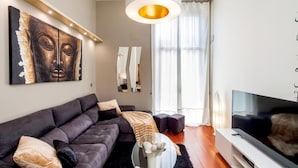 Una sala moderna con un sofá de color oscuro, un televisor de pantalla plana, una mesa redonda de centro y un cuadro abstracto grande en la pared.