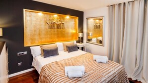 Una habitación de hotel con una cama grande, un cabecero con una obra de arte dorada, una mesita de noche con una lámpara y un espejo en la pared.