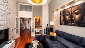 Una sala moderna con un sofá gris oscuro, una mesa de centro redonda de vidrio y un televisor sobre un mueble blanco. Se aprecia una gran obra de arte en la pared y una estantería con objetos decorativos.