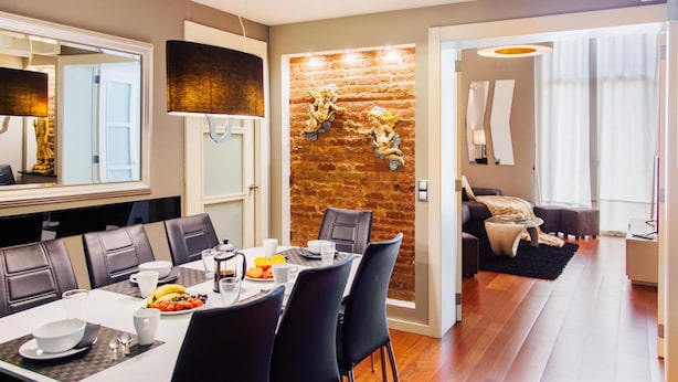 Un comedor moderno con una pared de ladrillo decorativa, un espejo grande y una zona de estar acogedora con sofá y reposapiés.