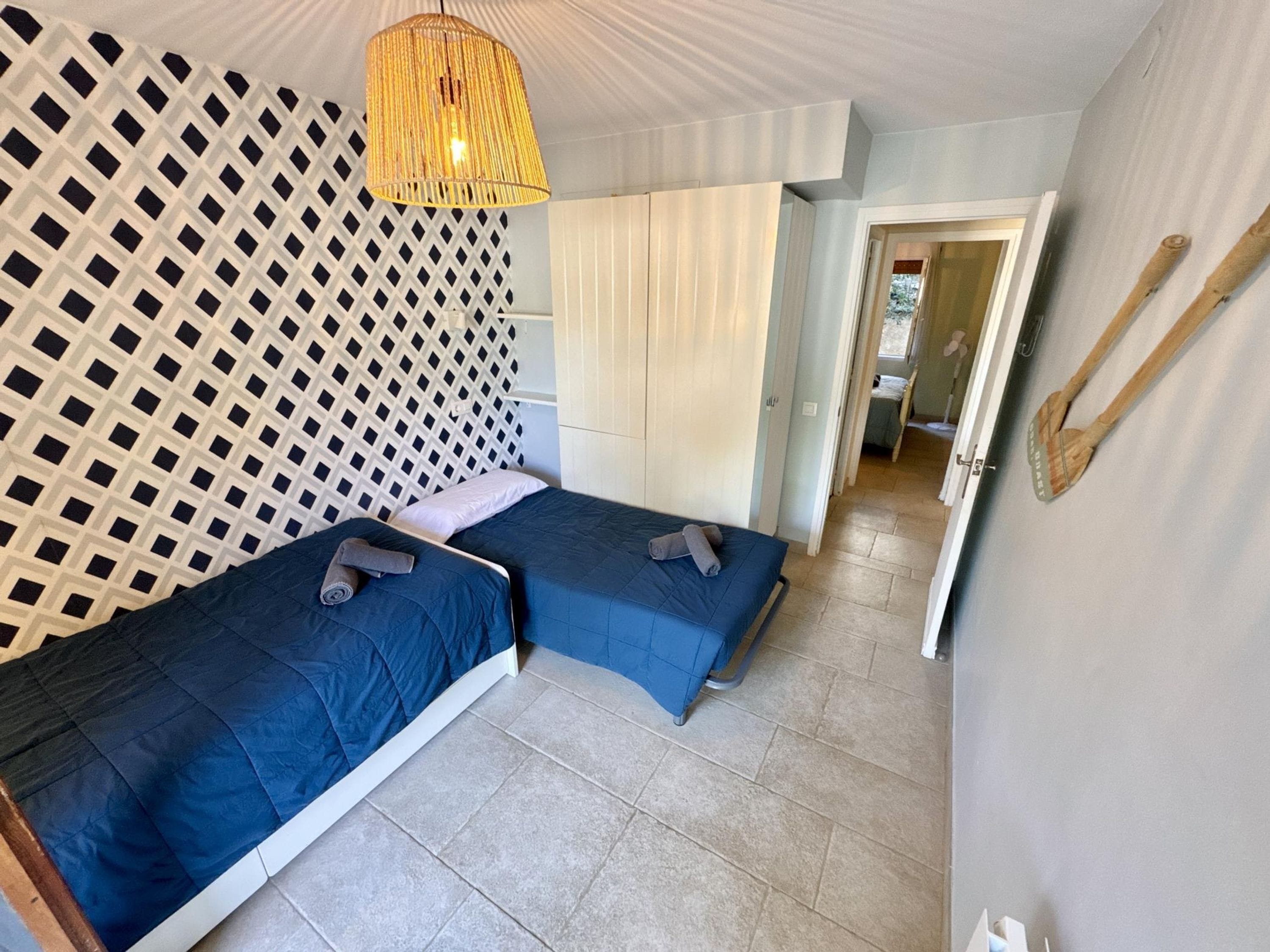 A bedroom with two beds, a patterned wall, a hanging light fixture, and a wooden spoon on the wall.