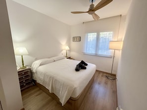 Ein Schlafzimmer mit einem Bett, zwei Nachttischen, einem Deckenventilator, einem Fenster mit Jalousien und einer Lampe auf jedem Tisch.