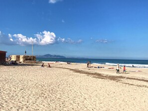 Une plage de sable avec une structure en pierre, des gens qui se détendent et une vue dégagée sur la mer et les montagnes lointaines.