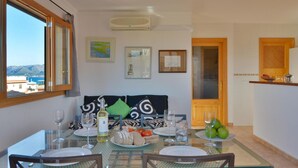 A dining area with a table set for four, a view of the sea, and a kitchen area with a fruit bowl.
