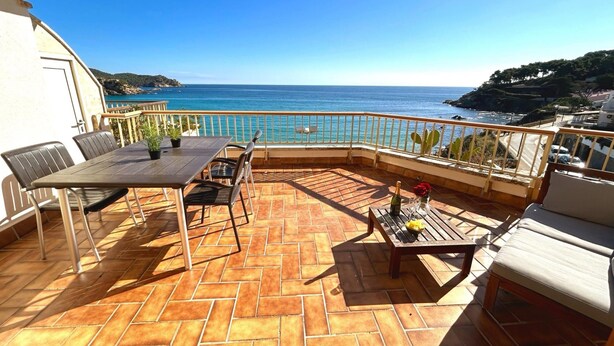 A terrace with a table and chairs, a small table with a bottle and a flower, and a view of the sea and hills. - Charming Apartment in La Fosca Beach (Palamós)