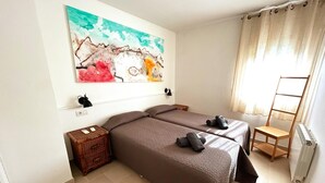 A room with two beds, a wooden nightstand, a radiator, and a painting on the wall. - Charming Apartment in La Fosca Beach (Palamós)