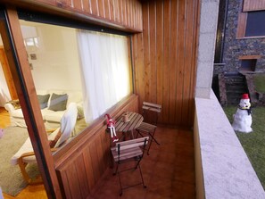 Una terraza de madera con un banco y una pequeña mesa, una puerta de cristal que da a una habitación con sofá y cortinas, y un edificio de piedra al fondo.