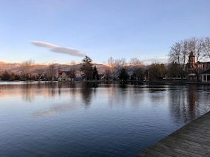 Un lago tranquilo con un muelle de madera, edificios y montañas al fondo.