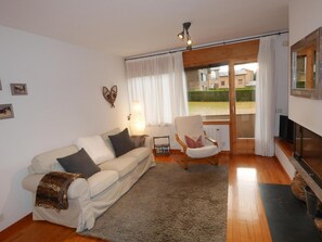 Una sala de estar con sofá, sillón y chimenea. Hay una ventana con cortinas, una lámpara y vistas al exterior.