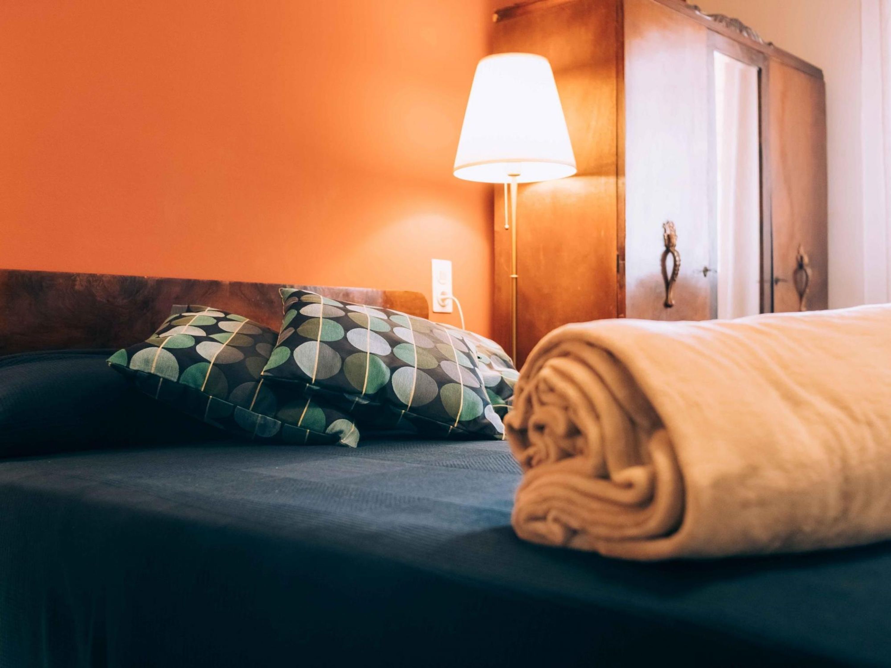 Un letto con una coperta beige arrotolata, due cuscini decorati e una lampada da comodino.