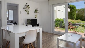A modern, well-lit room with a white desk, chairs, and a TV. There are two wall-mounted shelves with plants, and a sliding glass door leading to an outdoor patio area with a table and chairs. - Apartament amb Piscina i Parquing Baixos amb Jard (Palafrugell)