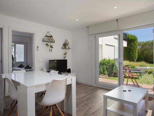 Una habitación moderna y bien iluminada con un escritorio blanco, sillas y un televisor. Hay dos estantes empotrados con plantas, y un ventanal corredero que da a un patio exterior con una mesa y sillas.