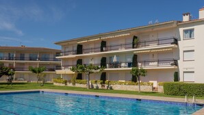 A swimming pool with a clear blue water, surrounded by a well-maintained lawn and a multi-story building with balconies. - Apartament amb Piscina i Parquing Baixos amb Jard (Palafrugell)