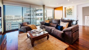 A modern living room with a sofa set, a coffee table, and a large window with blinds. - Victoria Diagonal Mar 2 Design Duplex Penthouse (Barcelona)